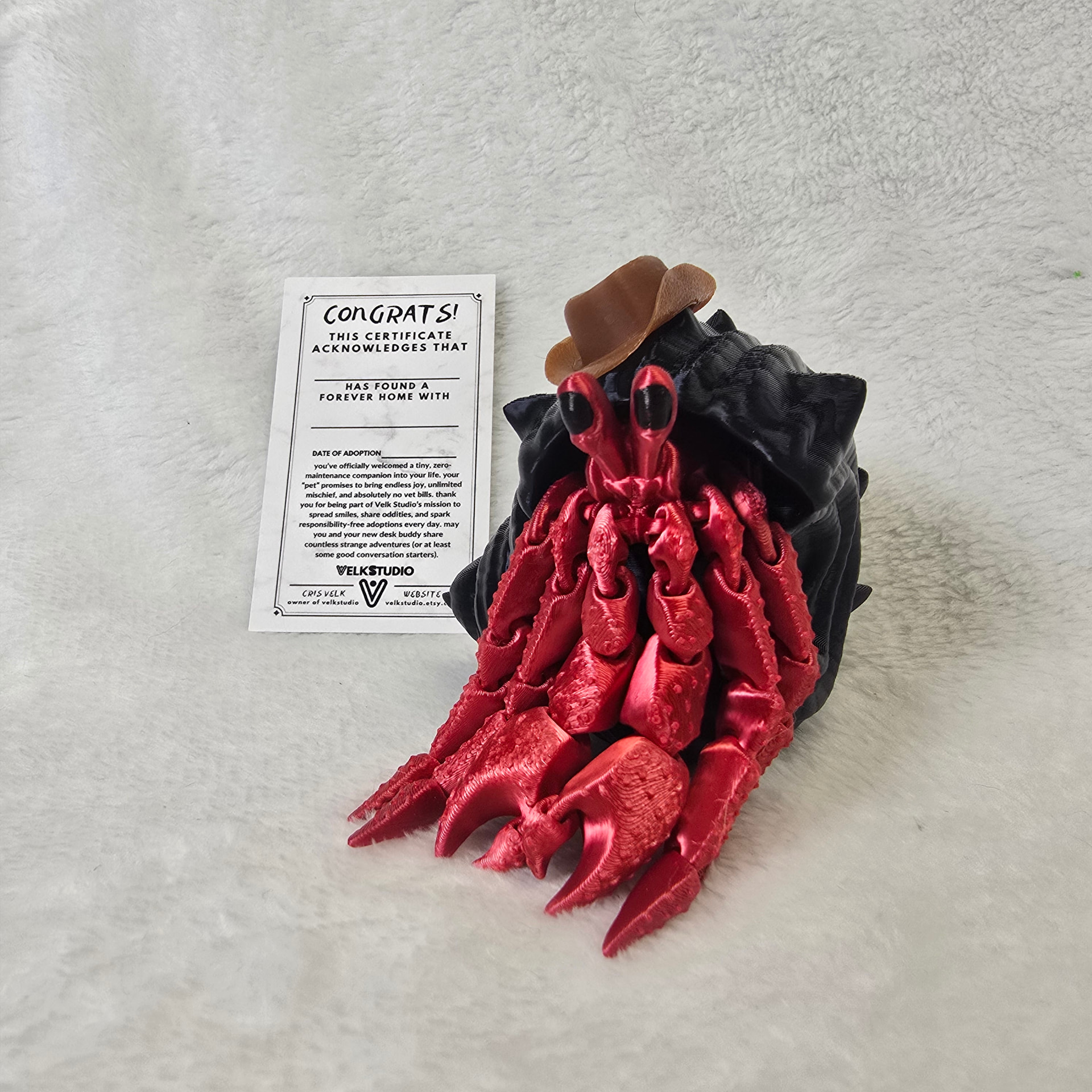 A red 3D printed hermit crab in a black shell wearing a brown cowboy hat, next to a Velk Studio adoption certificate